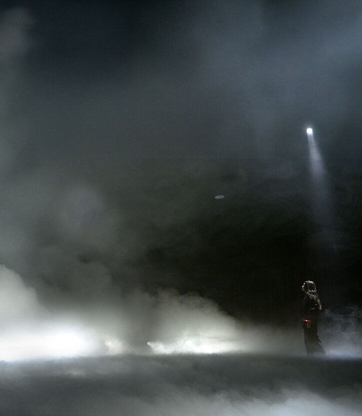 Eine Performerin steht vor einer riesigen Rauchwolke, die mit Licht bestrahlt wird.